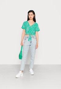 Top curto verde com padrão, com mangas curtas e laço na frente, combinado com jeans de perna reta de tom azul claro e ténis brancos de plataforma.