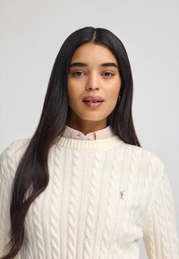 Kabelstickad krämfärgad tröja med hög hals, prydd med en liten broderad logotyp. Bärs över en rosa och vit rutmönstrad skjorta med krage.