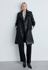 Schwarzer Leder-Trenchcoat mit doppelt gearbeiteten Knöpfen, großen Revers und seitlichen Taschen, kombiniert mit schwarzen maßgeschneiderten Hosen und Absatzstiefeln.