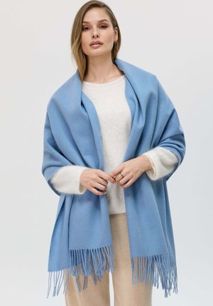 Scialle azzurro chiaro realizzato in morbido tessuto testurizzato con bordi frangiati. Indossato sopra un maglione beige, creando un look a strati.