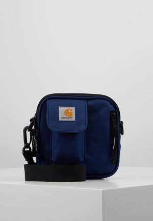 Mała granatowa torba Carhartt na ramię z przednią kieszenią na klapę, zamykanym na suwak przedziałem oraz regulowanym czarnym paskiem na białym tle.