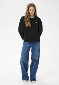 Schwarzer Pullover mit aufgepufften Ärmeln und kleinem weißem Herz-Logo auf der linken Brust, kombiniert mit weit geschnittenen blauen Denim-Jeans.