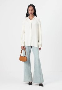 Camicia di seta bianca con maniche lunghe e ampie, abbinata a jeans a zampa d'elefante di colore azzurro chiaro. Gli accessori includono una piccola borsa a tracolla marrone e tacchi neri.