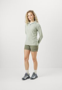 Hellgrüner Pullover mit langen Ärmeln und halbem Reißverschluss, kombiniert mit olivgrünen Shorts. Graue Wanderschuhe mit Schnürsenkeln und weißen Socken.