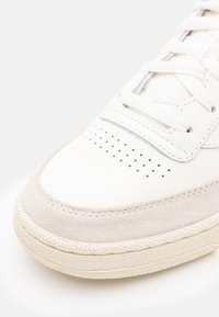 Vit lädersneaker med en tåkappa i mocka och perforerade detaljer. Har platta snören och en gummerad sula med struktur.