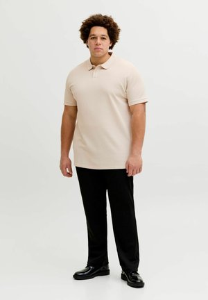 Mann mit lockigem Haar, der ein beiges Poloshirt, schwarze Hose und schwarze Lederschuhe trägt und vor einem schlichten Hintergrund steht.