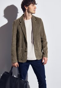 Blazer de pana color verde oliva con dos bolsillos frontales, cierre de un solo botón, llevado sobre una camisa clara, combinado con jeans oscuros y un bolso negro.