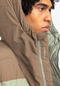 Quiksilver huva jacka i brunt och grönt, med en texturerad yta, vadderad foder och synlig dragkedja med logodetalj.