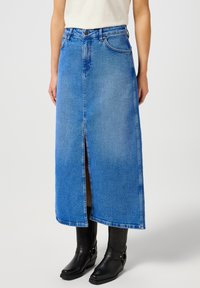 Heledsinine denim maxi seelik, millel on ees lõhik, klassikaline viie tasku disain ja vöökohal nupud, koos mustade sandaaliga saapatega.