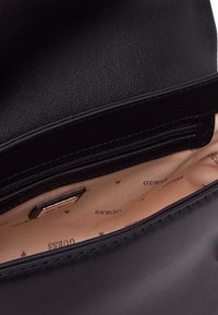 Interno di una borsa a tracolla in pelle nera con una fodera beige e piccoli loghi marroni. Include una tasca con zip per una sicurezza maggiore nello stoccaggio.