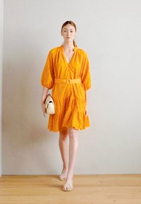 Kollane kleit puffvarrukatega, vöö ja kihilise hemiga. Hoiab valget kotti. Komplekteeritud nude toonides rihmkingadega, heledal taustal.
