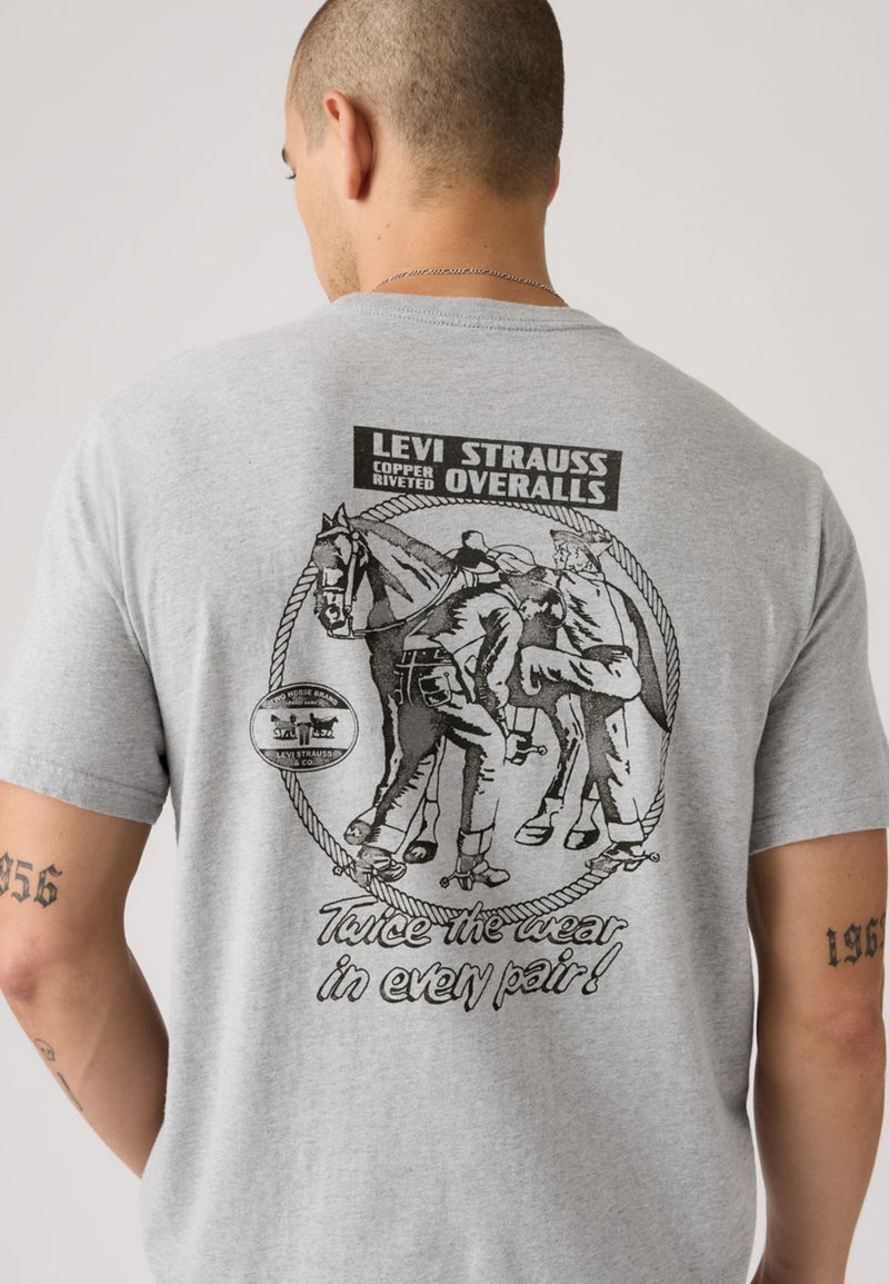 T-shirt en coton gris arborant un imprimé noir de deux personnages avec des chevaux et le texte « Levi Strauss Copper Riveted Overalls » au dos.