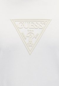Λευκό τριγωνικό λογότυπο με ανάγλυφο κείμενο «GUESS U.S.A.» και μεγάλο ερωτηματικό στο κέντρο σε λείο λευκό φόντο.