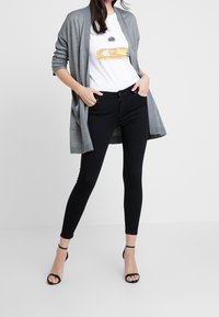 Šedý cardigan s otevřeným předním dílem, bílé tričko s potiskem, černé úzké džíny a černé sandály na podpatku se vzpěrami.