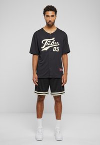 Svart baseballtrøye med en kremlogo og nummer "05", kombinert med svarte mesh-shorts og hvite joggesko med teksturerte lisser.