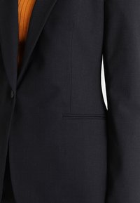 Mörkblå blazer med en slät textur, enfaldig knäppning och stela slag. Innehåller en diskret sidoficka och skräddarsydd passform.