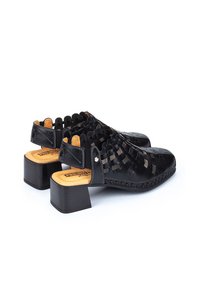 Chaussures à talons en cuir noir avec un dessus tissé, semelles en bois et talons carrés, accentuées de coutures et de petits détails en métal.
