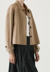 Cropped, beige wool jacket with a collar, three black buttons, and turned-up sleeves, paired with a white top and black skirt.