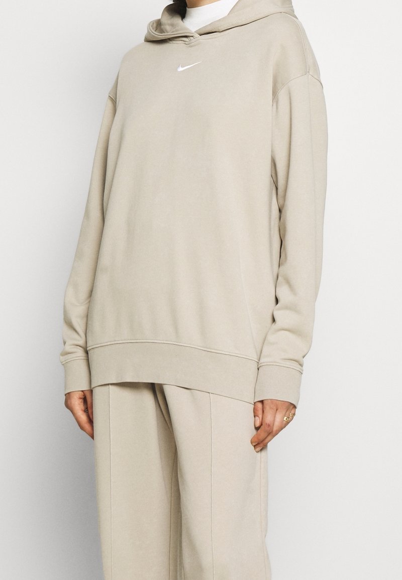 Ensemble sweat à capuche beige et pantalon assorti avec petit logo Nike blanc sur la poitrine, porté par une personne debout devant un fond uni.