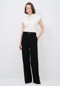 Weiße Bluse mit Schlüsselloch-Ausschnitt, kurzen Ärmeln; kombiniert mit schwarzen, hoch taillierten, weit geschnittenen Hosen; das Modell trägt goldene Akzente und einen Gürtel.