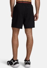Μαύρο αθλητικό σορτς από ελαφρύ υλικό με ελαστική μέση που φέρει κόκκινο κείμενο "UNDER ARMOUR", πάνω από γυμνά πόδια.