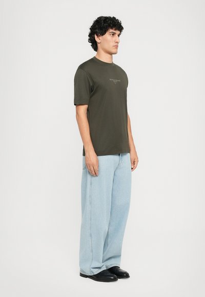 Fiatal férfi áll a fehér háttér előtt, olíva zöld, rövid ujjú pólót, világoskék, bő szárú farmert és fekete cipőt visel.