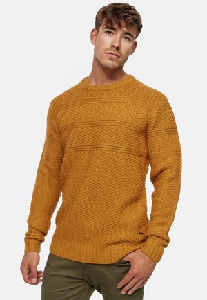 Senfgelber Strickpullover mit strukturiertem Muster, gerippten Bündchen und Saum. Crew-Neck-Design. Über olivgrünen Hosen getragen.