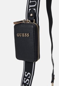 Černé kožené pouzdro na zip se zlatými kovovými doplňky. Má dlouhý popruh s bílým nápisem "GUESS". Kompaktní obdélníkový tvar.