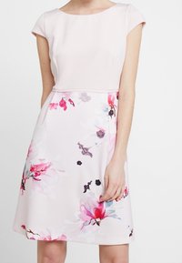 Donna che indossa un vestito rosa pallido con maniche a cappuccio e una gonna a stampa floreale con fiori rosa, rossi e viola.