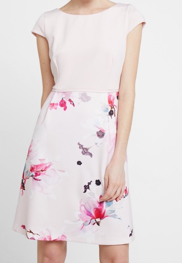 Donna che indossa un vestito rosa pallido con maniche a cappuccio e una gonna a stampa floreale con fiori rosa, rossi e viola.