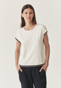 T-shirt bianco a maniche corte con scollo tondo, caratterizzato da accenti neri e beige lungo le maniche e l'orlo. Indossato con pantaloni neri.