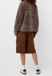 Chaqueta con estampado de leopardo en tonos marrón y beige, combinada con pantalones cortos cargo marrones. El conjunto presenta un ajuste relajado y bolsillos laterales visibles.