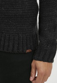 Suéter de punto gris oscuro con dobladillo y puños acanalados. Presenta una pequeña etiqueta marrón cerca del dobladillo. Patrón texturizado con motas de color.