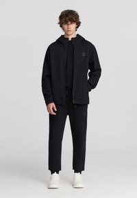 Felpa nera con zip e tasche frontali, abbinata a pantaloni della tuta neri con cordoncino. Entrambi realizzati in morbido tessuto, con accenti del logo discreti.