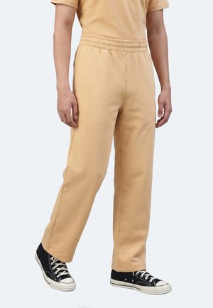 Persona che indossa pantaloni della tuta beige con vita elastica e sneaker nere basse con lacci bianchi, in piedi davanti a uno sfondo uniforme.