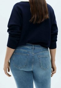 Femme portant un pull bleu marine et un jean bleu clair, vue de dos avec de longs cheveux bruns qui pendent.