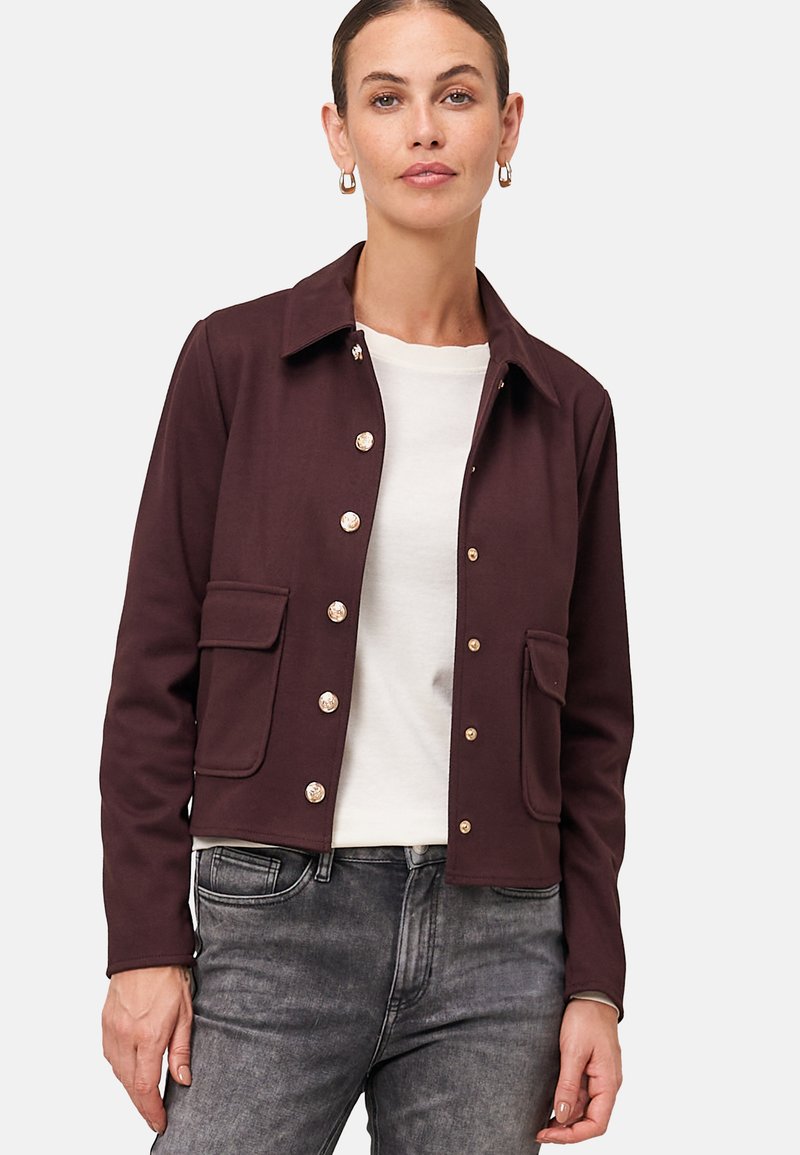 Veste courte bordeaux en tissu doux avec des boutons-pression métalliques, deux poches avant et un col. Chemise blanche en dessous.
