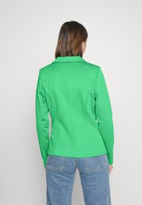 Zielona dopasowana marynarka wykonana z gładkiego materiału, z wyraźnie zaznaczonym kołnierzem i dopasowanymi szwami. W zestawieniu z jasnoniebieskimi spodniami dżinsowymi.