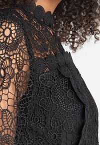 Tessuto in pizzo floreale nero sulla spalla e parte alta del petto di una donna, con dettagli in pizzo circolare e riccioli di capelli scuri parzialmente visibili.