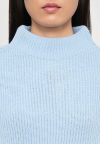 Gros plan sur le bas du visage et le cou d’une personne portant un pull en maille côtelée bleu clair à col montant.