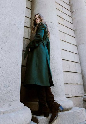 Femme en manteau long vert foncé et bottes brunes montant jusqu'aux genoux, se tenant entre de grandes colonnes en pierre, regardant vers le bas avec les mains dans les poches du manteau.