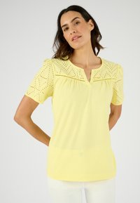 Korte mouwen gele top met een v-hals, voorzien van eyelet-details op de schouders en borst, en een losse pasvorm. Zachte, lichte stof.