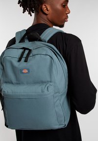 Mochila de lona azul claro con dos correas para los hombros, bolsillo frontal con cremallera y una insignia con logo. Textura suave y forma rectangular con costuras reforzadas.