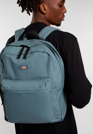 Zaino in tela azzurro chiaro con due spallacci, tasca frontale con zip e badge con logo. Texture liscia e forma rettangolare con cuciture rinforzate.