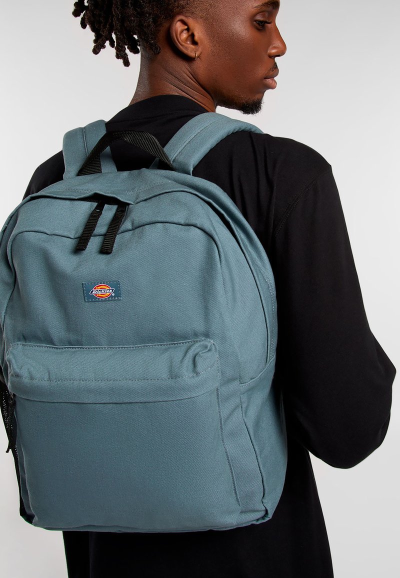Mochila de lona azul claro con dos correas para los hombros, bolsillo frontal con cremallera y una insignia con logo. Textura suave y forma rectangular con costuras reforzadas.