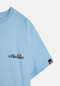 Gaiši zila kokvilnas T-kreklu ar melnu "ellesse" logotipu un krāsainu emblemu kreisajā krūtī. Īsas piedurknes, apaļa kakla izgriezums un sānu logotipa detaļa.