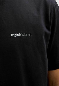 Schwarzes Baumwoll-T-Shirt mit einem weißen, gestickten Logo, das auf der linken Brustseite "Originals™ STUDIO" liest. Glatte Textur.