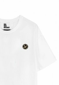 Camiseta blanca de algodón con cuello redondo y mangas cortas, que presenta un logotipo bordado circular en negro y dorado en el pecho.