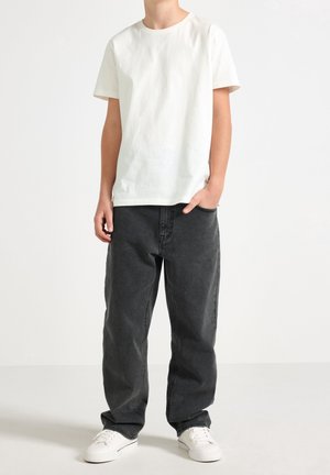 Person wearing a plain white short-sleeve t-shirt, loose dark gray jeans, white sneakers, standing with one hand in pocket against a white background.