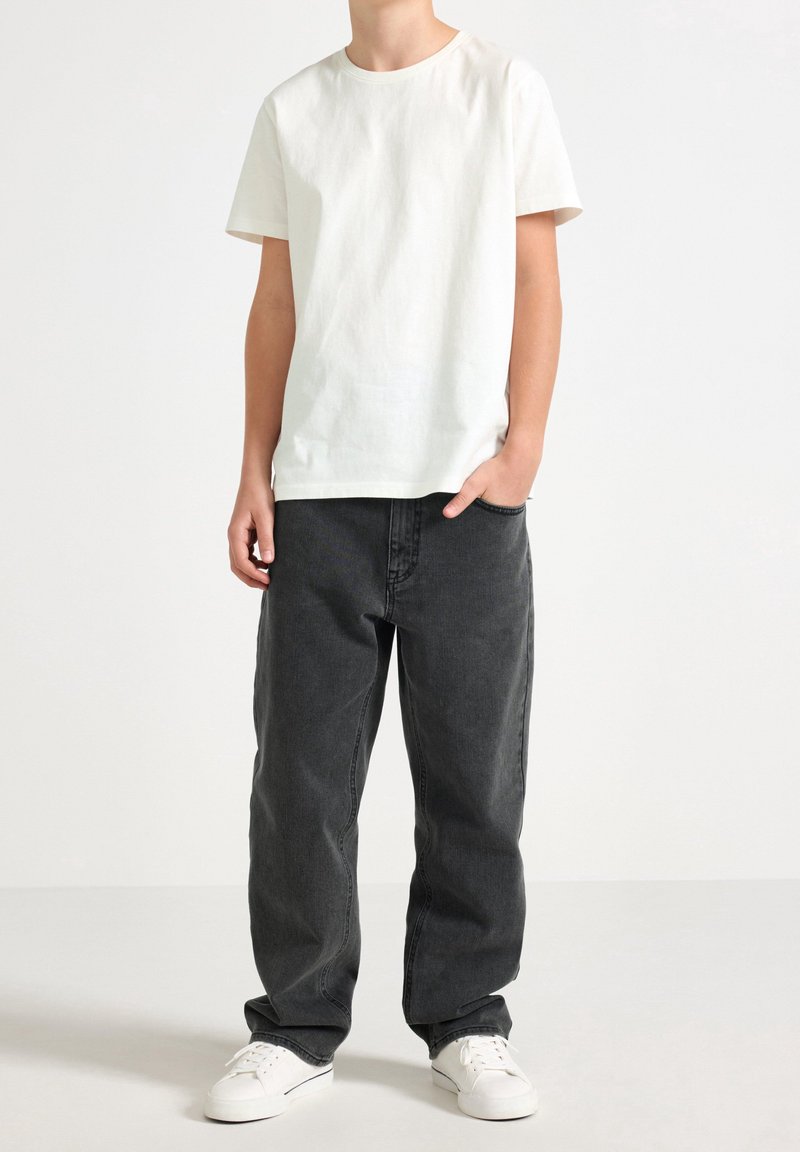 Personne portant un t-shirt blanc uni à manches courtes, un jean gris foncé large, des baskets blanches, debout avec une main dans la poche sur un fond blanc.